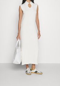 Abito a maglia bianco senza maniche, lunghezza al ginocchio, con scollatura a chiave. Abbinato a sneakers grigie con strisce nere e una borsa a tracolla bianca.