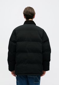 Chaqueta acolchada negra con cuello alto, que presenta un diseño acolchado y un ajuste holgado. La tela parece suave con un acabado mate.