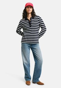 Marineblauwe en witte gestreepte blouse met lange mouwen en een met knopen sluitende kraag, gecombineerd met lichtblauwe jeans en bruine enkellaarzen, afgemaakt met een rode pet.