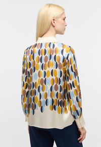Blusa di seta con un design a motivo che presenta cerchi sovrapposti in giallo, marrone, blu e crema. Polsini con bottoni e colletto classico.