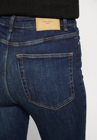 Vista trasera de una persona que lleva jeans de mezclilla azul oscuro con un bolsillo trasero visible y una etiqueta de cuero marrón de la marca Vero Moda.