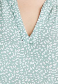 Light blue blouse featuring a V-neckline, adorned with a white leaf pattern, made from a soft, textured fabric.