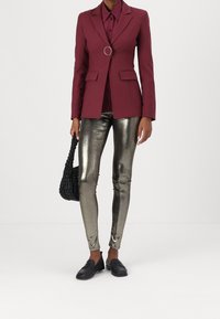 Blazer marrón rojizo con un detalle redondo, combinado con una camisa burdeos oscuro, leggings plateados metálicos y mocasines negros, sosteniendo un bolso negro con textura.