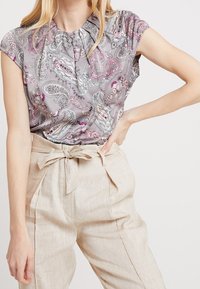 Blouse grise avec un motif cachemire en tons roses et bleus, manches courtes, encolure froncée ; associée à un pantalon beige taille haute.