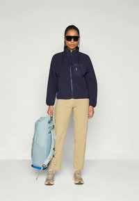 Γυναίκα που φοράει σκούρο μπλε ζακέτα με φερμουάρ, μπεζ παντελόνι, μπεζ αθλητικά παπούτσια και μεγάλα μαύρα γυαλιά ηλίου, κρατώντας μια ανοιχτό μπλε σακίδιο στο πλάι της.