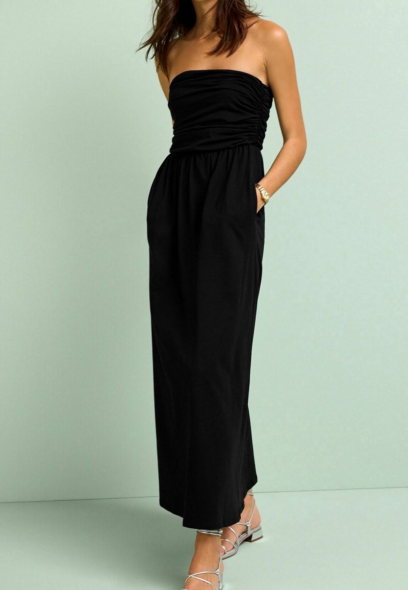 Robe maxi noire sans bretelles avec un corsage froncé, des poches sur les côtés et une jupe fluide. Portée avec des sandales argentées à lanières sur un fond vert menthe.