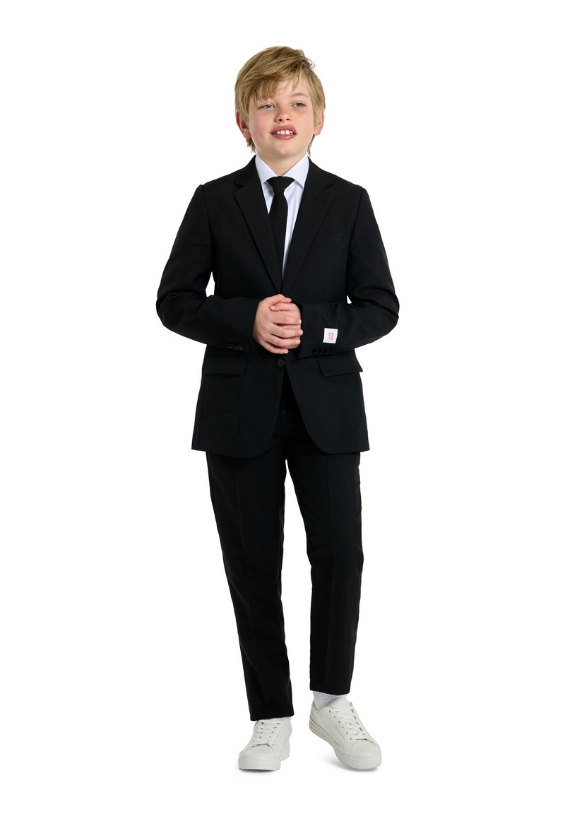 Czarny garnitur skrojony na miarę z białą koszulą i czarnym krawatem. Białe sneakersy kontrastują z formalnym ubraniem. Krótkie blond włosy stylizowane na luzie.