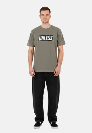 Uomo in piedi che indossa una maglietta verde oliva con la scritta "UNLESS", pantaloni neri larghi e scarpe slip-on nere su uno sfondo bianco semplice.