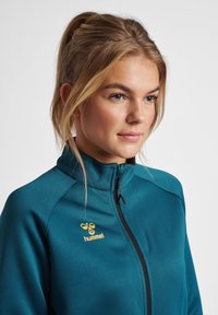 Zielona kurtka zapinana na zamek, wykonana z miękkiego, strukturalnego materiału, z wysokim kołnierzem i żółtym logo na lewej piersi.