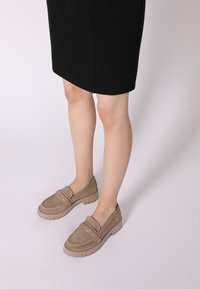 Beige suède instappers met een dikke rubberen zool en decoratieve stiksels. Gecombineerd met een zwarte rok, waarbij de ronde neus en het instapontwerp goed tot hun recht komen.