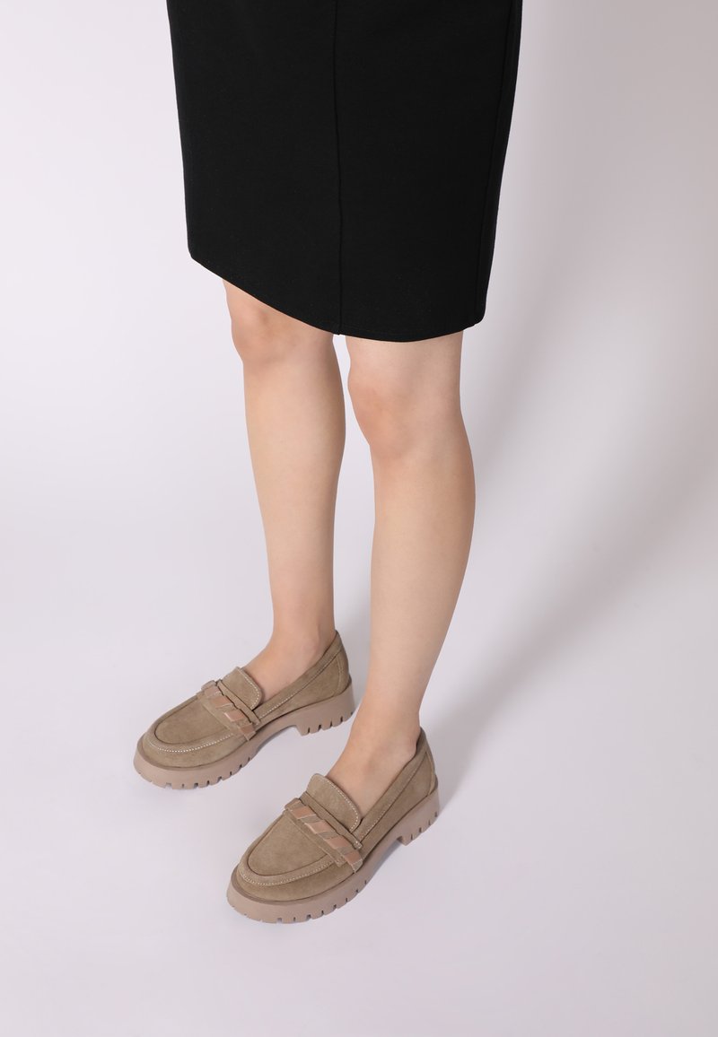 Beige suède instappers met een dikke rubberen zool en decoratieve stiksels. Gecombineerd met een zwarte rok, waarbij de ronde neus en het instapontwerp goed tot hun recht komen.