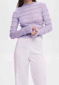 Personne portant un haut à manches longues lavande texturé et un pantalon blanc taille haute, les mains jointes près de la taille.