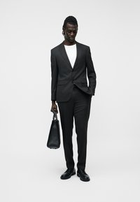 Άνδρας που φοράει μαύρο κοστούμι και λευκό πουκάμισο, κρατώντας μια μαύρη δερμάτινη τσάντα, στέκεται μπροστά σε ένα απλό φωτεινό φόντο.