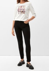 Weißes Langarmhemd mit einem bunten Textgrafik, kombiniert mit schwarzen Slim-Fit-Jeans und schwarzen Slippern mit dekorativem Kettendetail.
