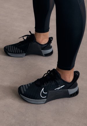 Pieds portant des chaussures de sport noires avec un logo blanc et des leggings noirs debout sur un sol beige.
