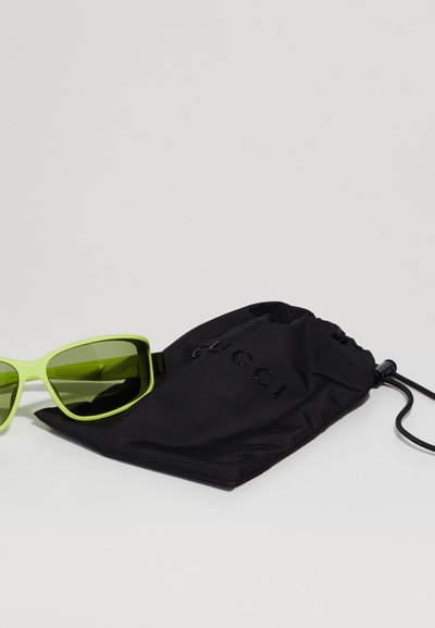 Gafas de sol rectangulares verdes con montura gruesa junto a un estuche de tela negra con texto en relieve. El estuche tiene un cierre de cordón.