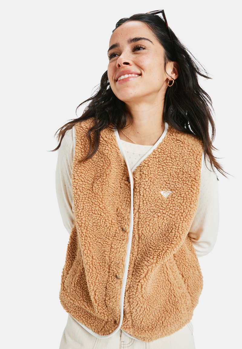 Beige Fleece-Weste mit strukturierten Oberflächen, weißen Akzenten und Druckknopfverschlüssen vorne. Sie verfügt über ein kleines Logo auf der linken Seite.