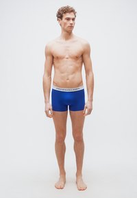 Νέος άνδρας που στέκεται ξυπόλητος φορώντας μπλε boxer briefs Polo Ralph Lauren με λευκή ζώνη στη μέση.