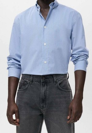 Mężczyzna w jasnoniebieskiej koszuli zapinanej na guziki z podwiniętymi rękawami, włożonej do wysokich, czarnych jeansów z wyblakłym efektem.