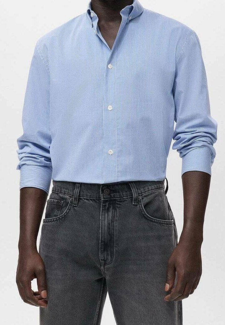 Homme portant une chemise bleue claire à boutons avec manches retroussées, rentrée dans un jean noir délavé taille haute.