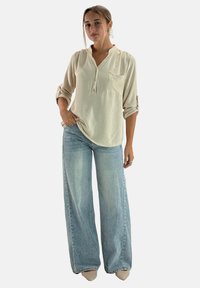 Jasno beżowa koszula na guziki z jedną kieszenią na piersi, w połączeniu z szerokimi, wyblakłymi niebieskimi dżinsami, zaprezentowana na modelce.