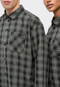 Fekete-fehér kockás gombos ing, elterjedt gallérral, mellzsebbel és kontrasztos kockás mintával. Puha anyag textúra.