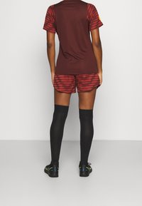 Bordeauxfarbenes Sportshirt mit gemusterten Ärmeln, passende bordeauxfarbene Shorts und schwarze Kniestrümpfe. Schwarze Sneakers mit neonfarbenen Akzenten.