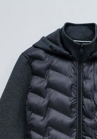 Gilet rembourré avec un devant noir brillant et des manches et une capuche en tissu texturé gris charbon. Il dispose d'une fermeture éclair à l'avant et d'une capuche à cordon.