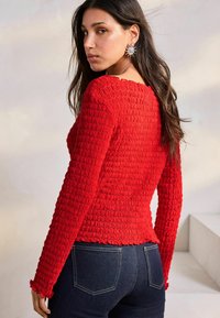 Haut long à manches rouges texturé avec une silhouette ajustée et un ourlet festonné, associé à un jean foncé. Le tissu présente un motif smocké.