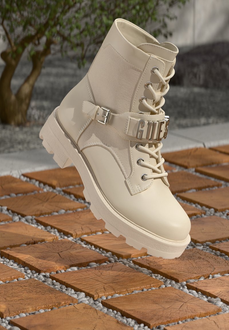 Bota de tornozelo em marfim com acabamento texturizado, tiras de fivela prateada, design de atacador e sola robusta, posicionada acima de um caminho de pedra.