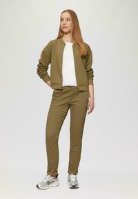 Braune strukturierte Bomberjacke über einem weißen T-Shirt, kombiniert mit olivgrünen, schmal geschnittenen Hosen und silbernen Sportschuhen.