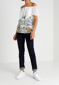 Blouse blanche décolleté des épaules avec des détails en volants, ornée d'imprimés floraux colorés et de paysages urbains, associée à un jean roulé noir et des baskets blanches.