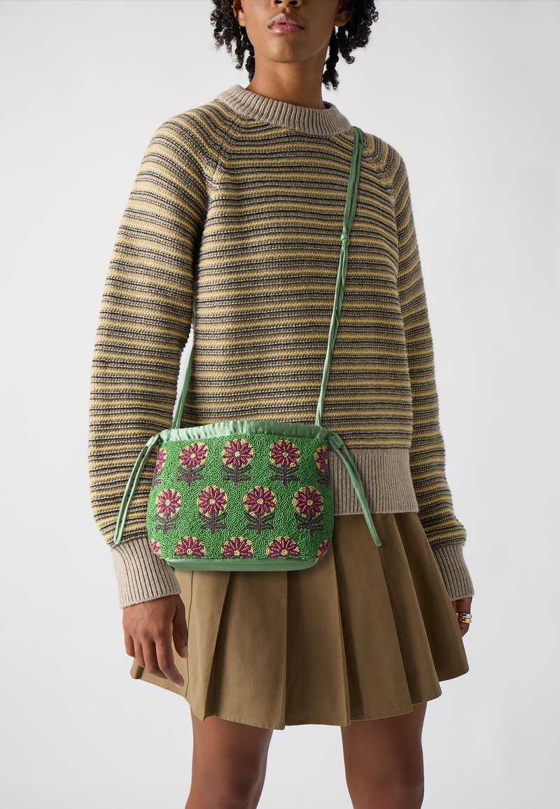 Grüne florale Umhängetasche mit strukturiertem Oberflächen, die rosa Blumen und grüne Stiele zeigt. Lange, dünne Träger. Getragen mit einem gestreiften Pullover und einem Rock.