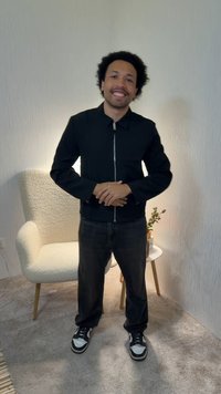 Άνδρας με κατσαρά μαλλιά που φοράει μαύρο σακάκι και παντελόνι, στέκεται και χαμογελά σε ένα δωμάτιο με μοκέτα, λευκοί υφασμάτινοι τοίχοι, λευκή καρέκλα και τραπεζάκι με φυτό.