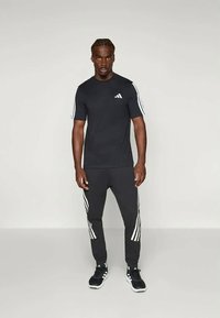 Črna atletska majica z belim Adidas logotipom in striped detajli, v kombinaciji s črnimi jogger hlačami z belimi črtami ob straneh.