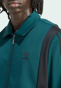 Teal zip-up jakke lavet af glat stof, med en sort stribe, ribbet krave og broderet sort Adidas-logo på brystet.