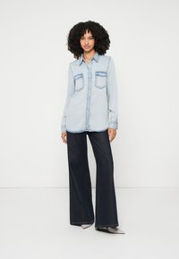 Light blue denim shirt with two chest pockets, button-up front, and long sleeves, paired with dark wide-leg jeans and pointed shoes.