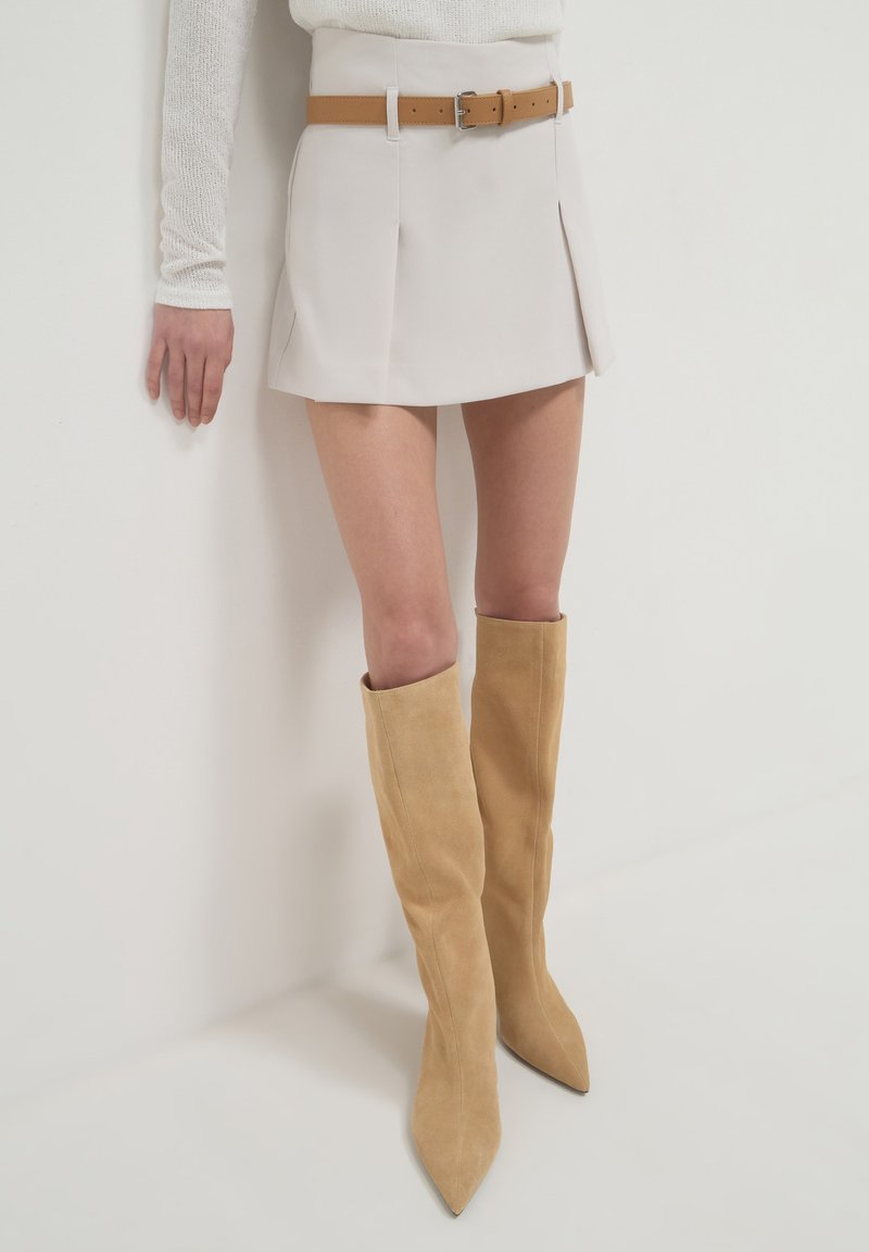 Beige Faltenminirock mit einem braunen Leder-Gürtel, kombiniert mit knielangen, sandfarbenen Wildlederstiefeln mit spitzen Zehen.