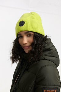 Neon zelená rebrová čiapka s kruhovou čiernou nášivkou. Nosená s tmavozelenou páperovou bundou s nastaviteľnou kapucňou a šnúrkami.