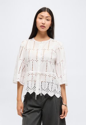 Jonge vrouw met lang donker haar draagt een witte blouse met oogjes, knopen en ceintuur in de taille, gecombineerd met donkere, wijde broekspijpen.