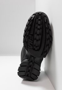 Mustan urheilukengän pohja, jossa on syvä ja teksturoitu kuvio, kestävä kumimateriaali ja logo, joka on painettu pohjaan pidon parantamiseksi.