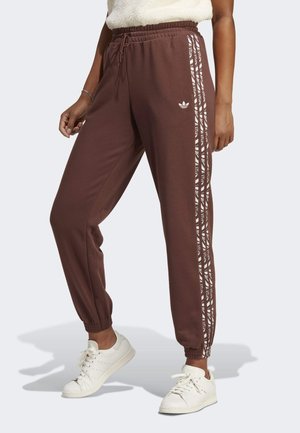 Person wearing brown Adidas track pants with white patterned side stripes and white sneakers, standing with one foot slightly forward.