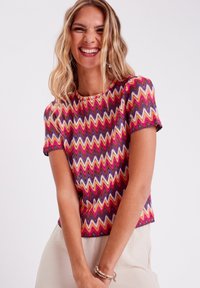 T-shirt à manches courtes avec un motif zigzag multicolore, présentant des teintes vives de rose, violet et orange. Texture lisse et col rond.