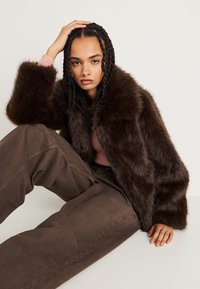 Casaco curto de pêlo sintético castanho com um grande colarinho, usado sobre uma blusa rosa, combinado com calças castanhas largas. Aparência texturizada e suave.