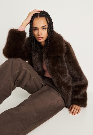 Casaco curto de pêlo sintético castanho com um grande colarinho, usado sobre uma blusa rosa, combinado com calças castanhas largas. Aparência texturizada e suave.