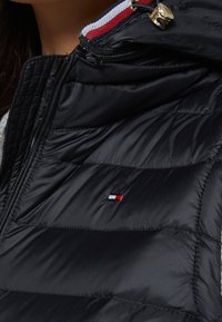 Svart puffärmad väst med horisontella quiltade sektioner, dragkedja fram och en liten broderad logotyp i rött, vitt och blått på bröstet.