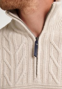 Maglione beige a maglia con colletto alto, caratterizzato da un motivo a treccia e una zip argento con un pomello blu.