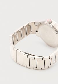 Ασημένιο μεταλλικό μπρασελέ ρολογιού με κούμπωμα χαραγμένο με την επιγραφή «HUGO», προσαρμοσμένο στο πίσω μέρος στρογγυλής κάσας ρολογιού με αυλακωτό κορώνα.