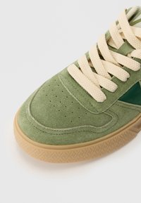 Grøn ruskinds sneaker med cremefarvede snørebånd, perforeringer på tåboksen og en gummigummisål. Siden har et grønt accentpanel.