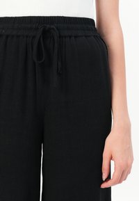 Zwarte, loszittende broek met een elastische tailleband en trekkoorddetails. De stof lijkt zacht te zijn met een gladde textuur.
